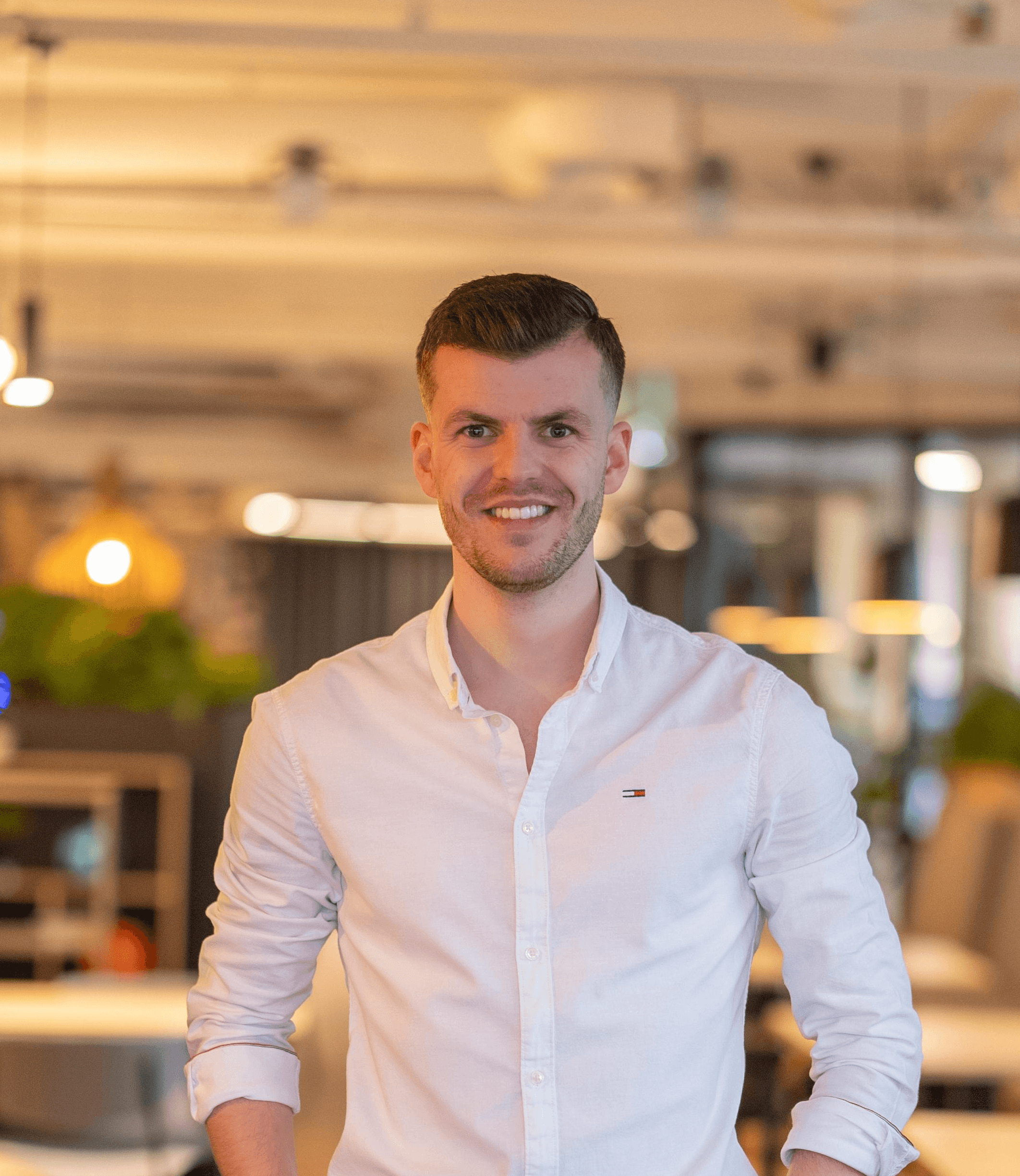 Young man in a black turtleneck and blazer stands outdoors, smiling softly. The background is blurred, conveying a warm, professional tone.