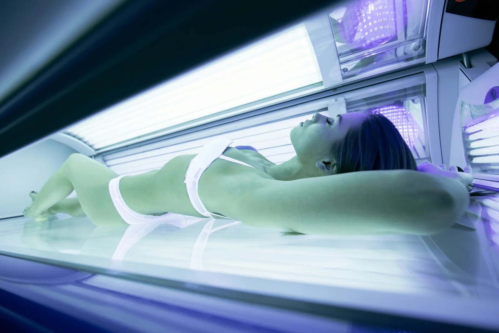 Person lying in tanning bed with UV lights illuminated during indoor tanning session.