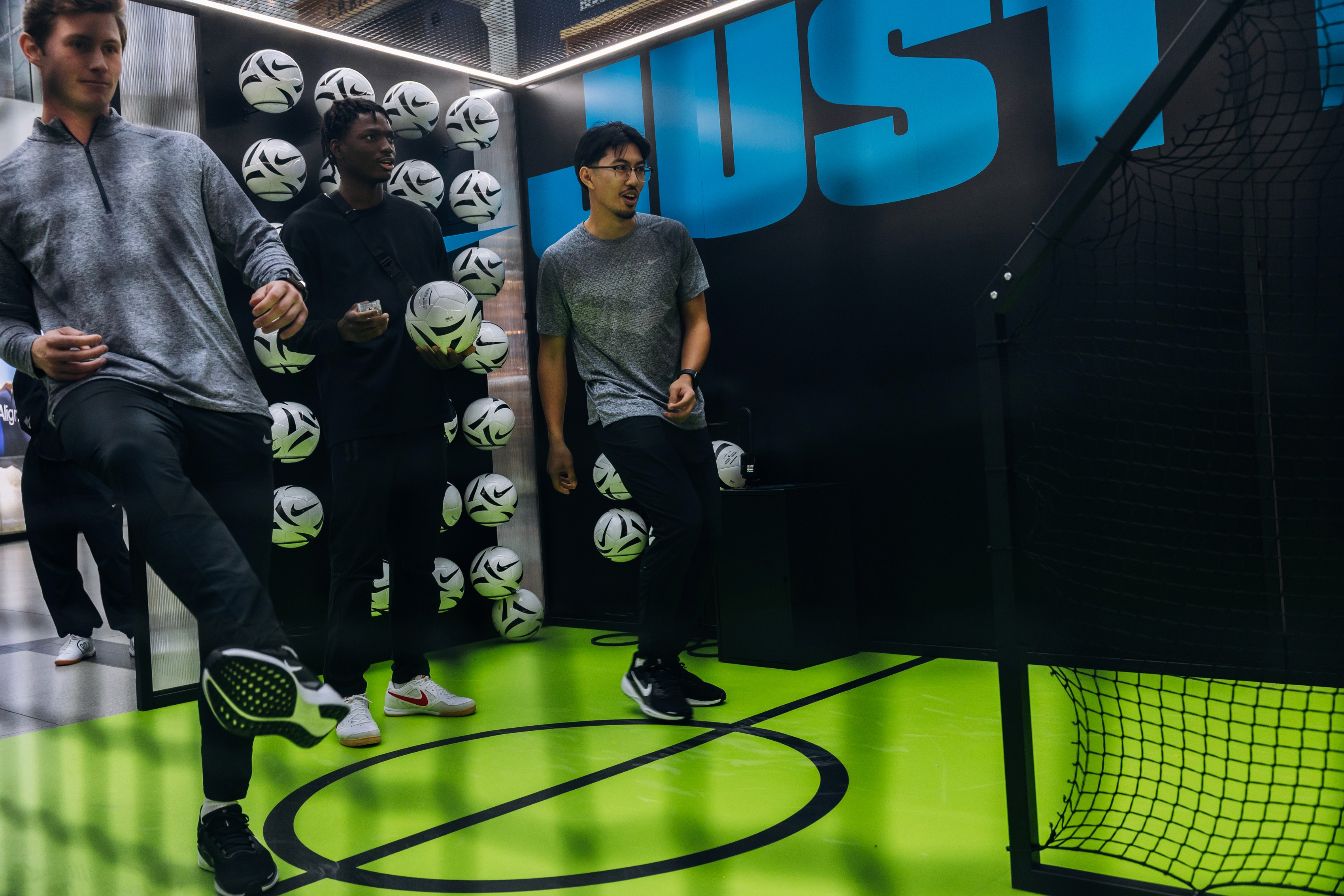 Three people in casual sportswear smile and kick soccer balls in a modern, neon-green sports zone, with walls adorned with soccer balls and bold text.