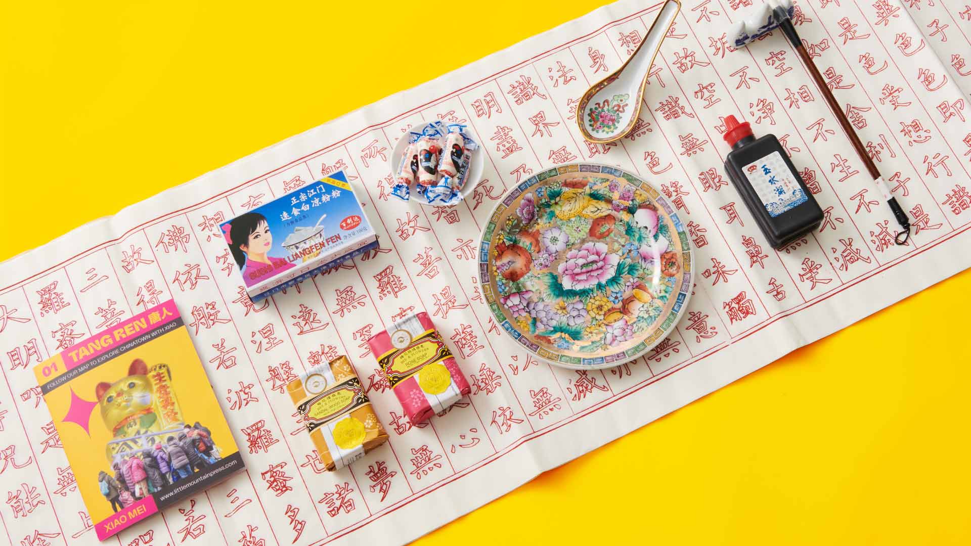 A yellow background features a white cloth with red Chinese characters and symbols. On the cloth are several items: a yellow box with a cartoon cat image and Chinese text, a blue box with Chinese characters, a small white bowl with red and blue candies, a decorative plate with floral designs, a small black bottle with Chinese characters, and a white spoon. A red and white striped stick is visible near the top right corner.