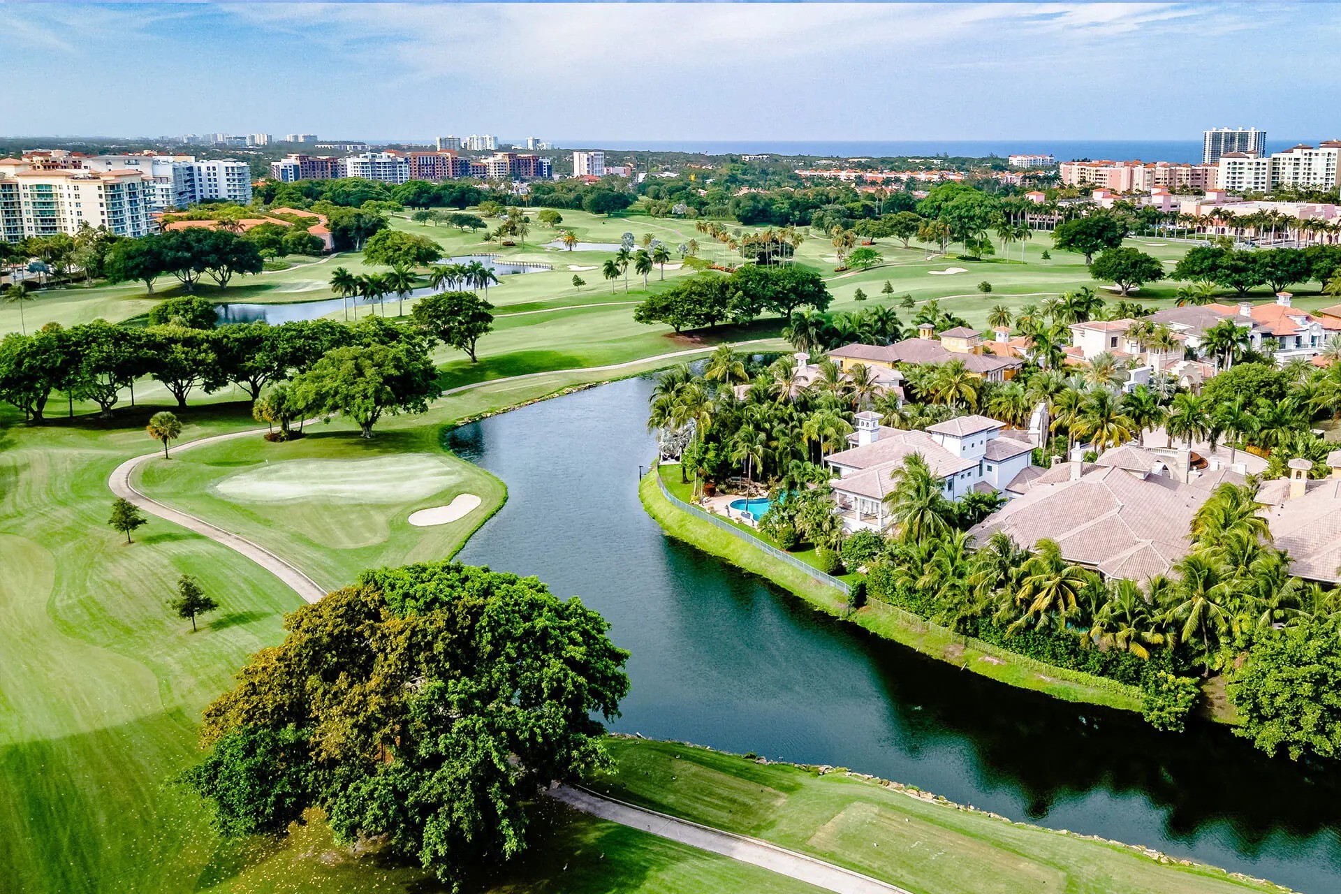 A breathtaking aerial view of a lush golf course in Boca Raton, Florida, featuring manicured greens, winding water features, and luxurious waterfront estates surrounded by palm trees. In the background, upscale residential buildings and the ocean skyline complete the picturesque scene.