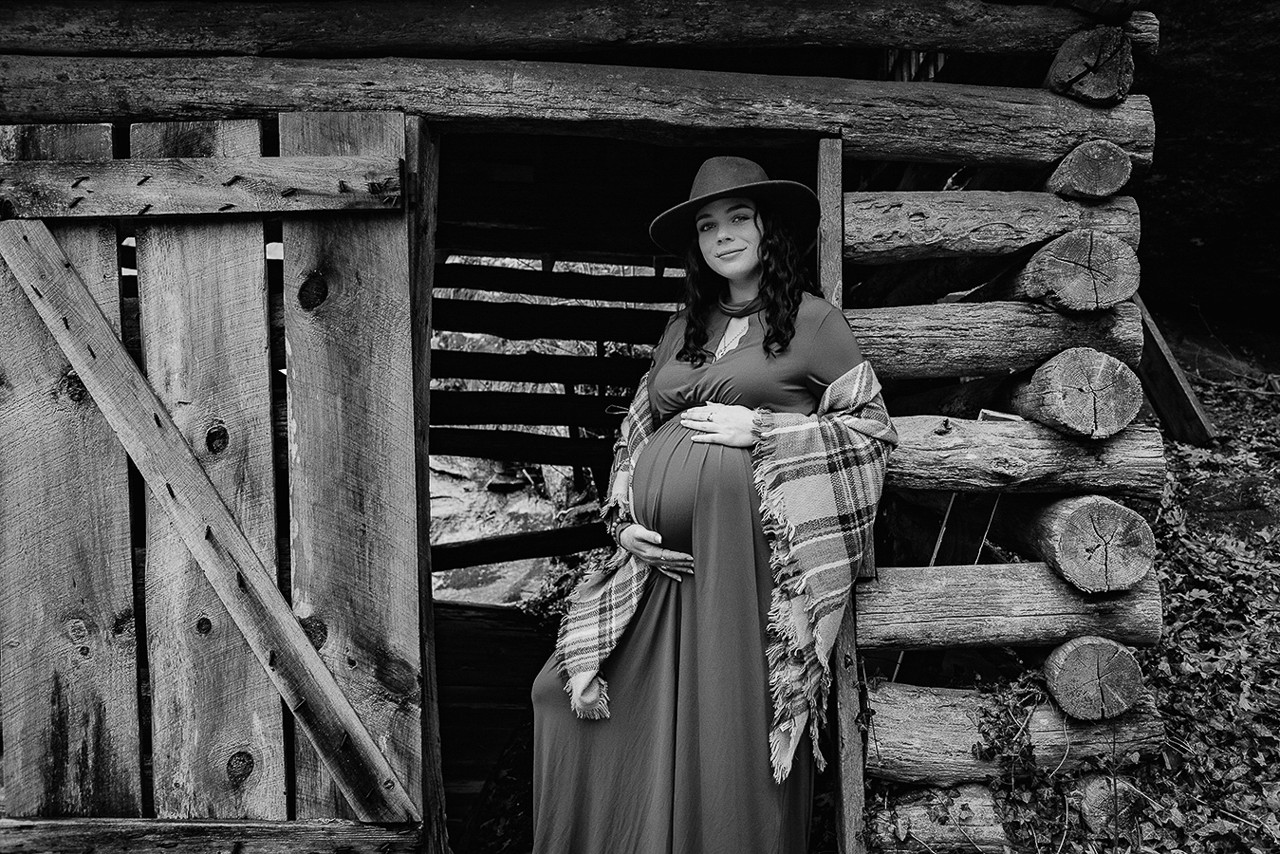Pregnant woman in red dress and hat, standing in a wooden doorway, hands on stomach. Maternity photo.