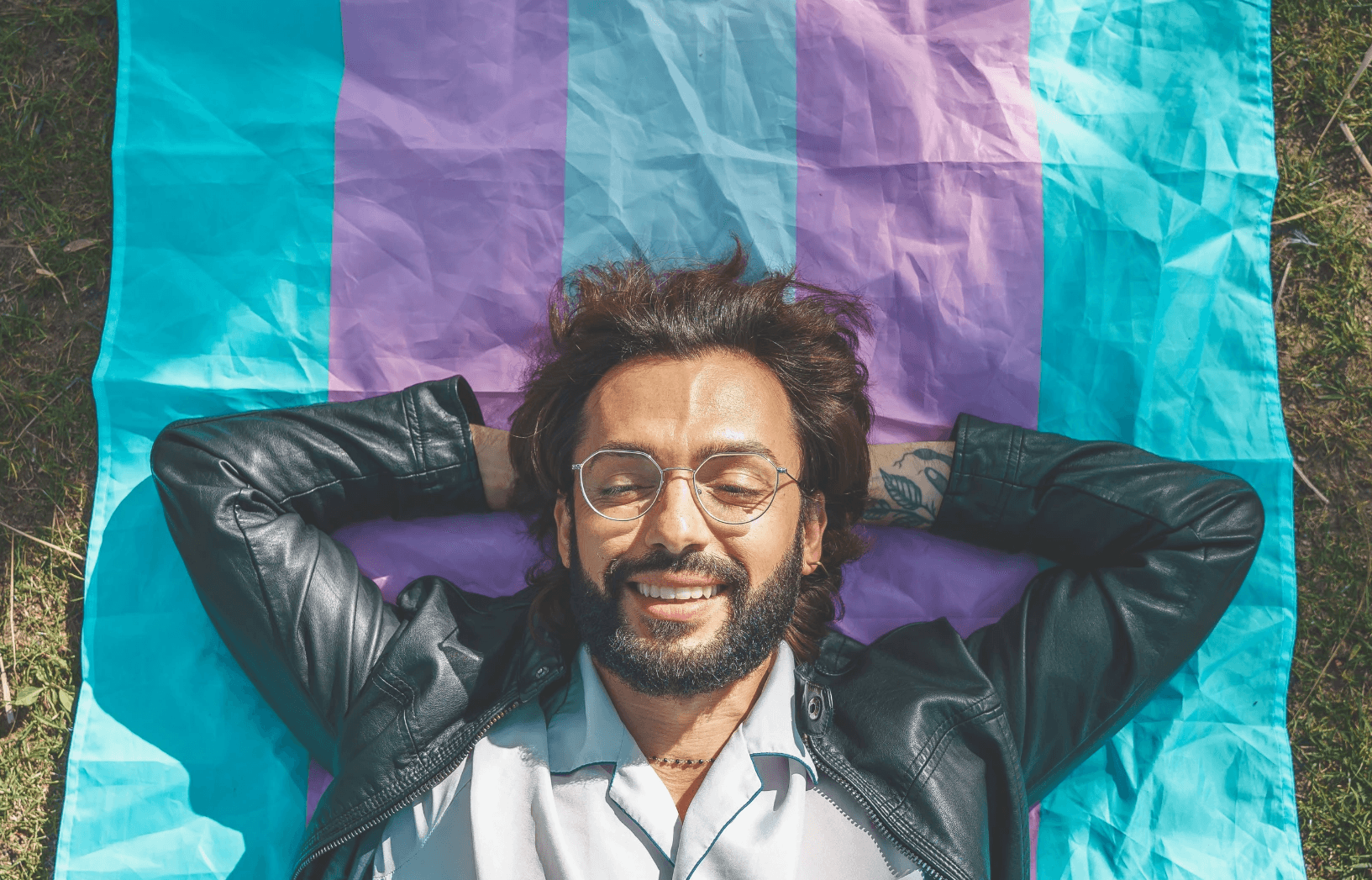 A smiling man with glasses lies on a colorful blanket, enjoying the outdoors. Green grass surrounds him.