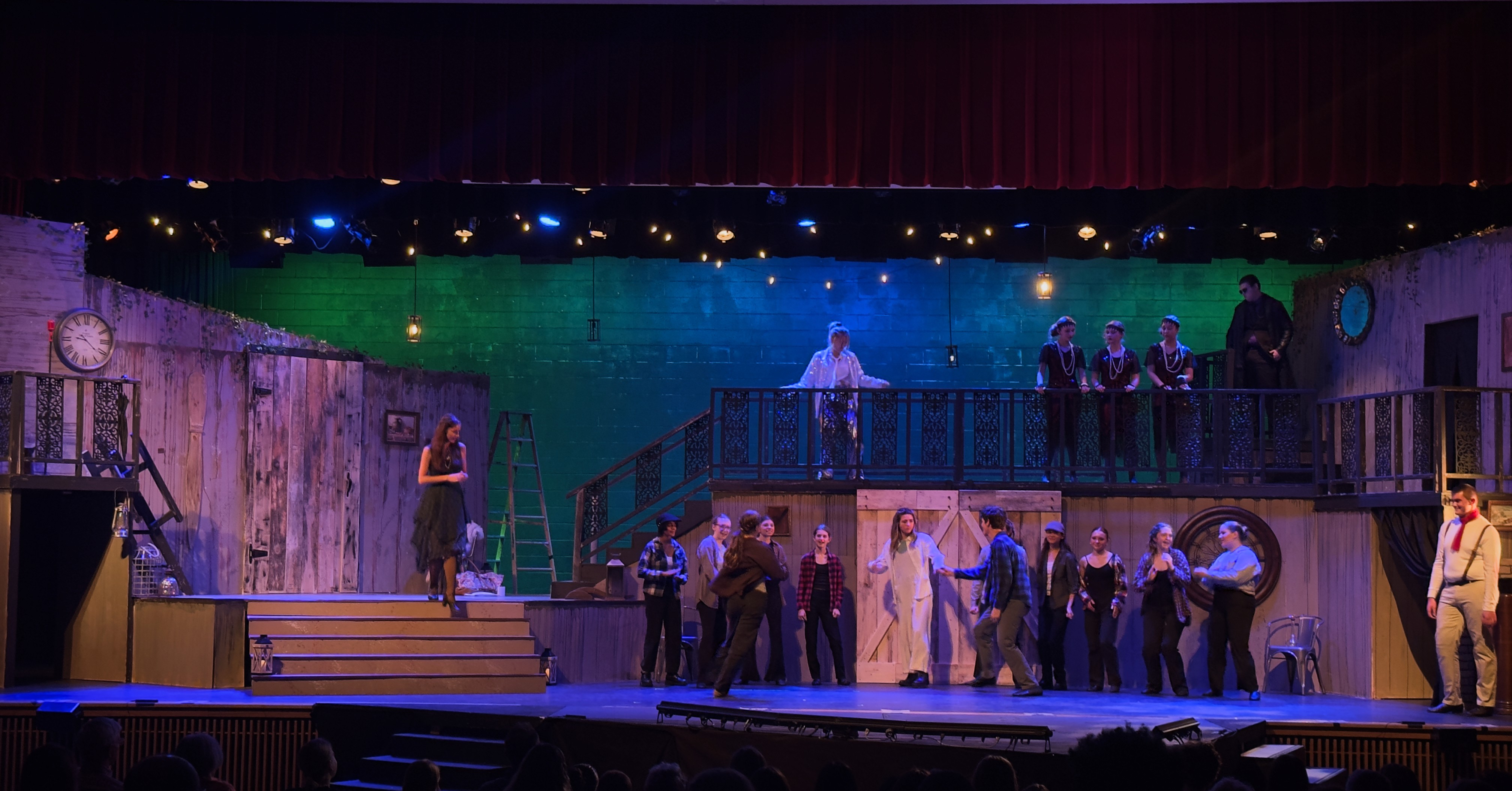 A vibrant stage set features a green and blue gradient lighting backdrop with performers engaging in a subtle dance number, enhanced by warm amber side lighting.