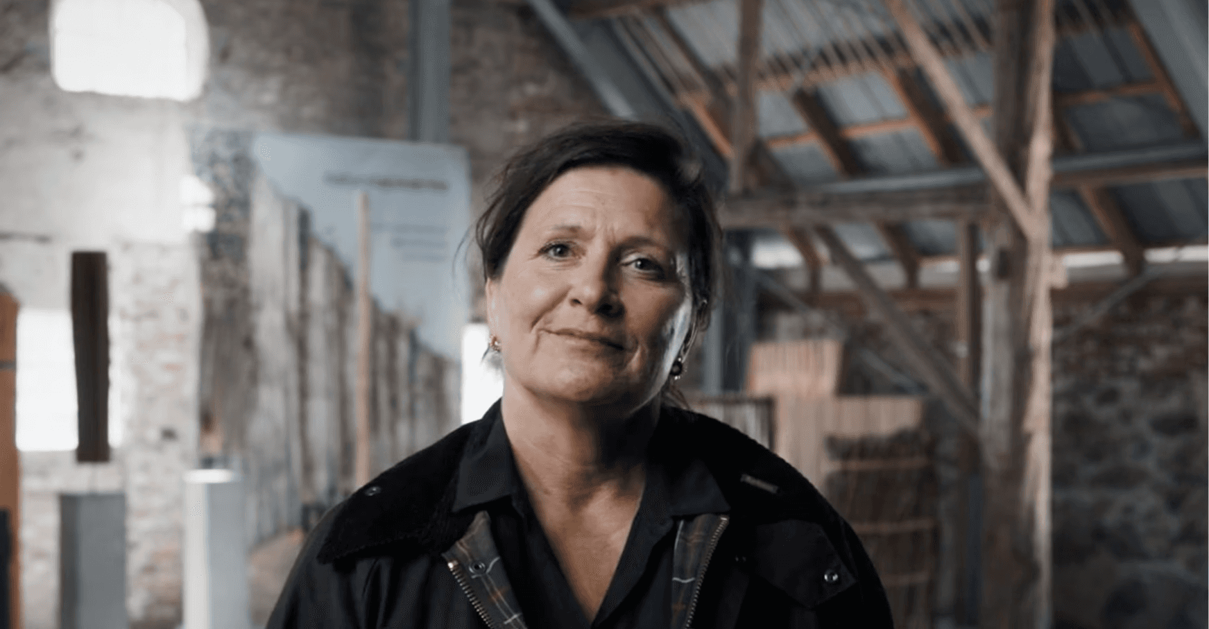 Woman with brown hair and brown jacket, smiling in front of inner farmhouse.