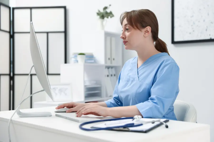 Nurse in blue scrubs using AI medical software on a desktop computer at a bright clinical workstation.