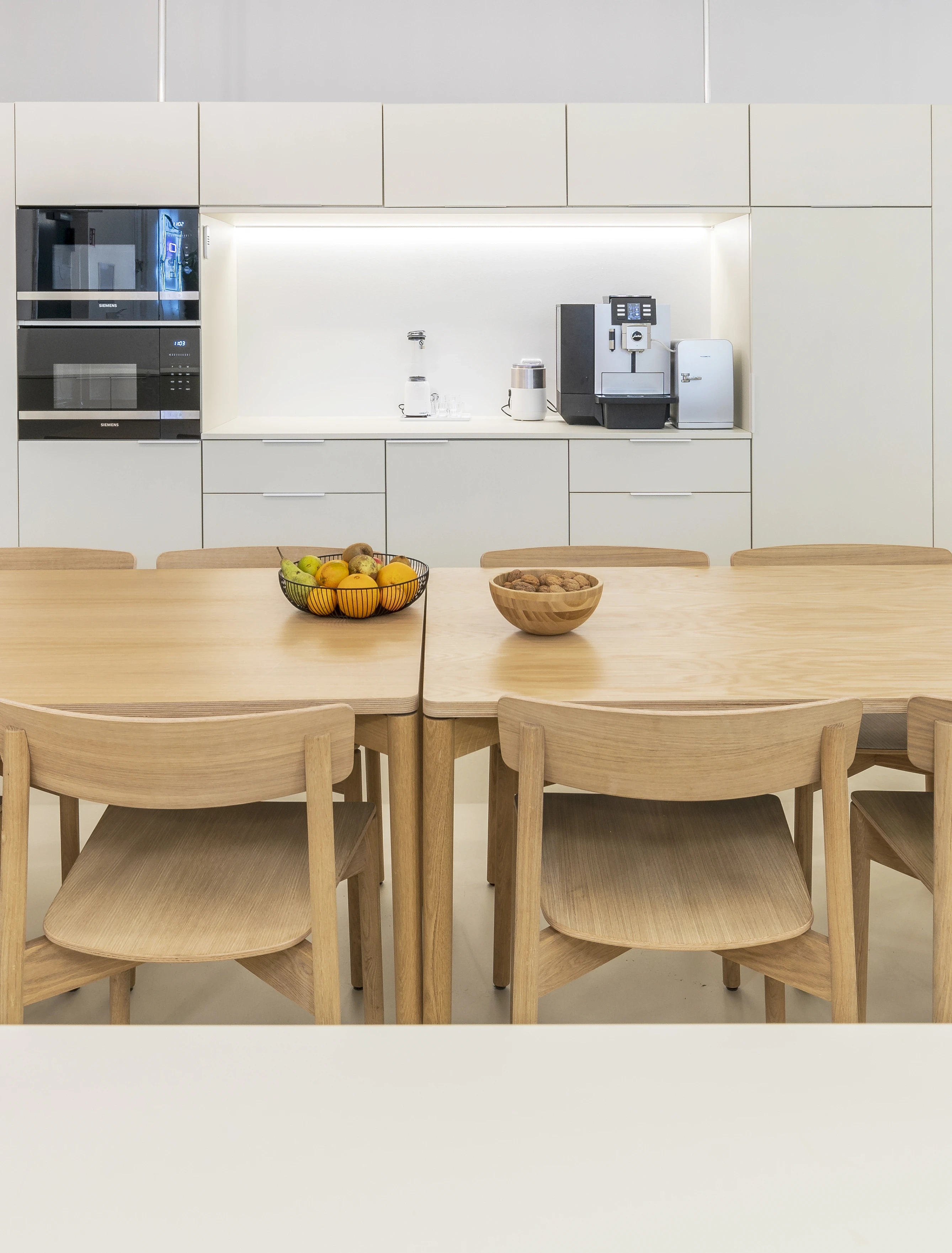 Kitchen at ELEMENT with flat white cabinets, built-in appliances and coffee machines, fruit bowls on two pale oak tables with matching wooden chairs.
