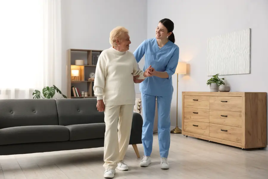 Home caregiver in blue scrubs assisting an elderly woman with walking across a bright living room.