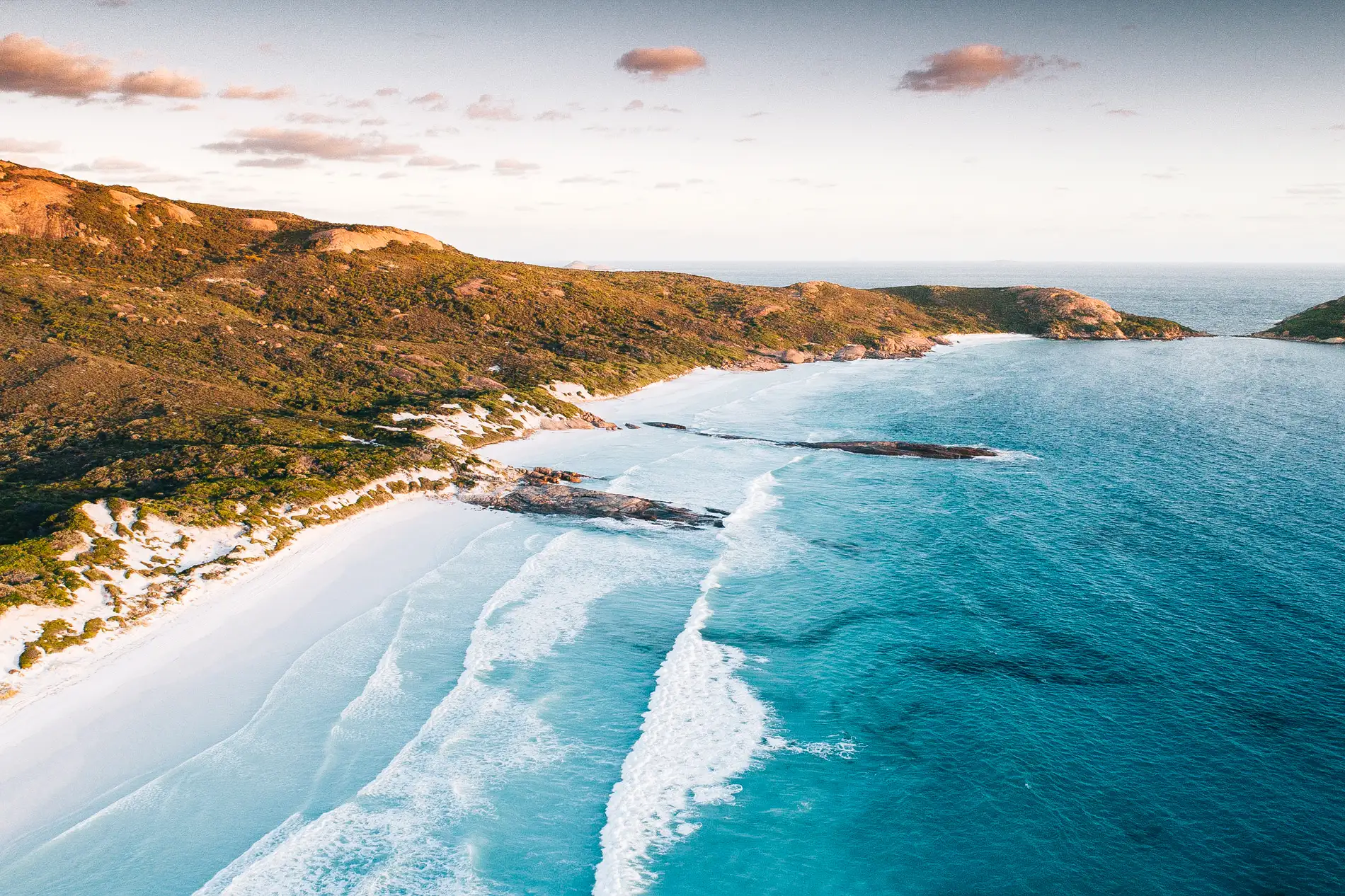 Aerial of surf breaking onto a white beach below a golden headland at dusk