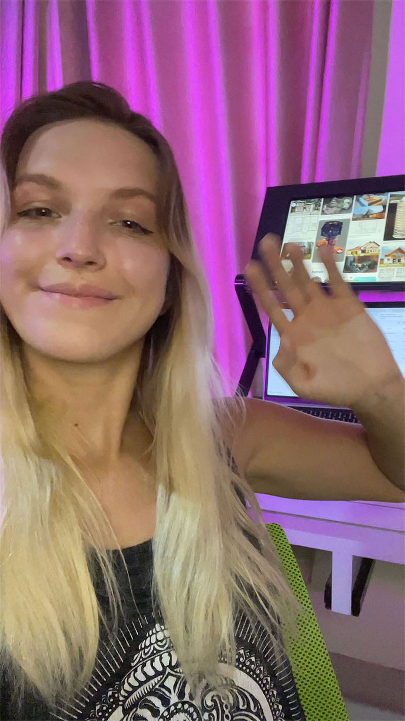 A person with long blonde hair smiles and waves in front of a purple background, holding a tablet.