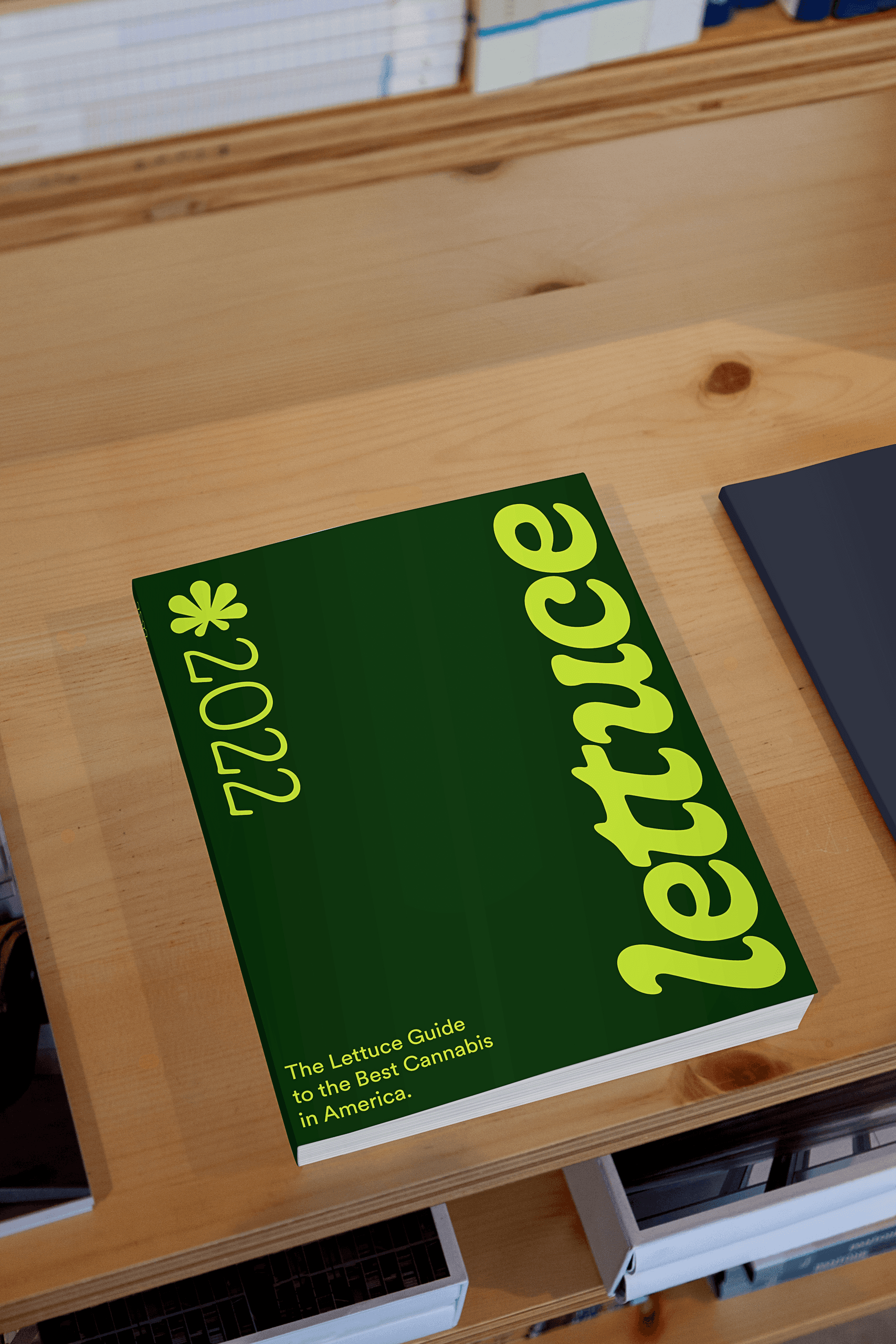 A dark green book titled “Lettuce 2022” rests on a light wooden surface. The title is written in lime green with a stylized font. The subtitle, “The Lettuce Guide to the Best Cannabis in America,” is printed in smaller white text below the title. A dark object is partially visible to the right of the book, and a window with blinds is visible in the background.