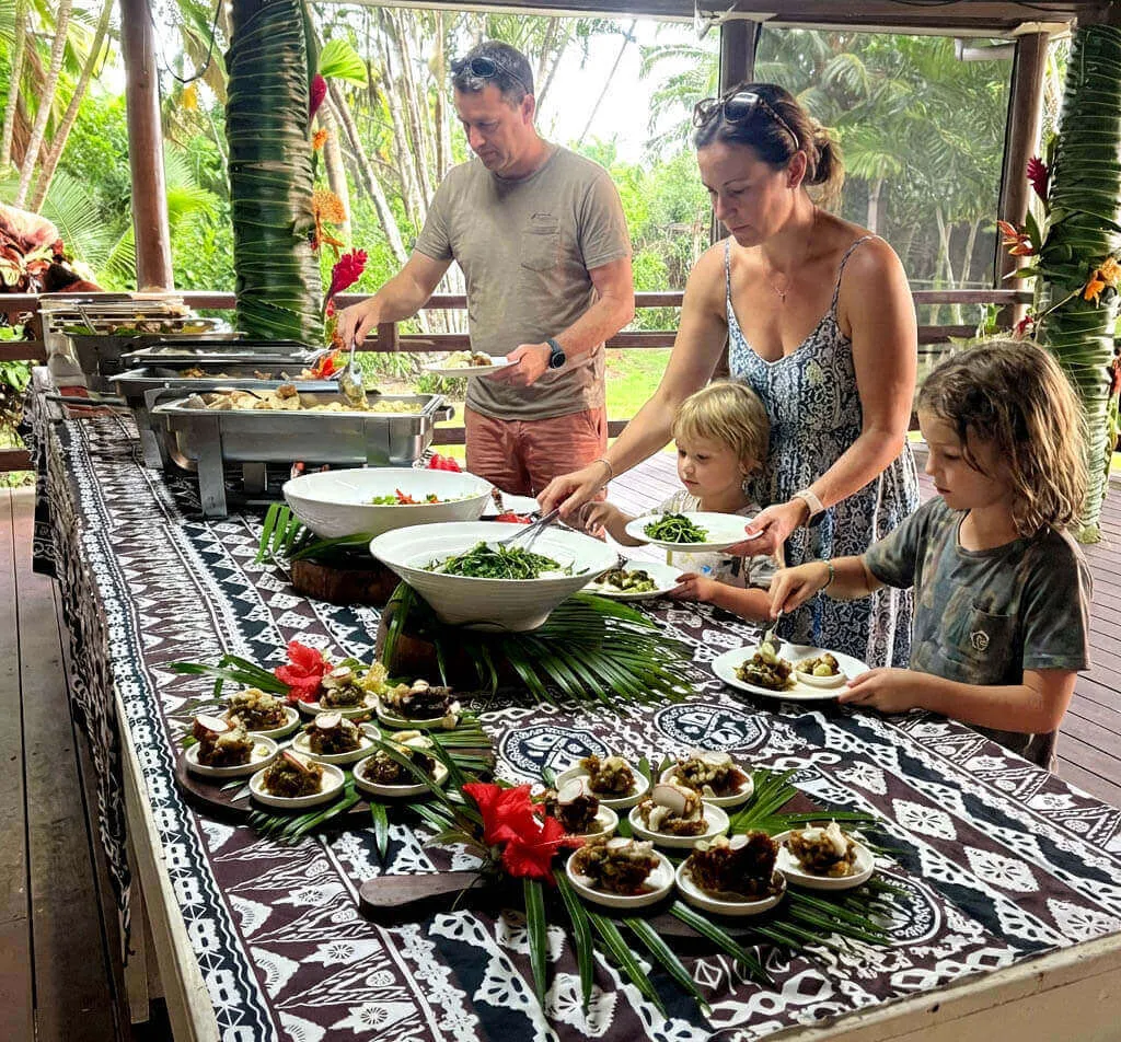 Family serves themselves at a buffet table with traditional Fijian decorations in a tropical resort.