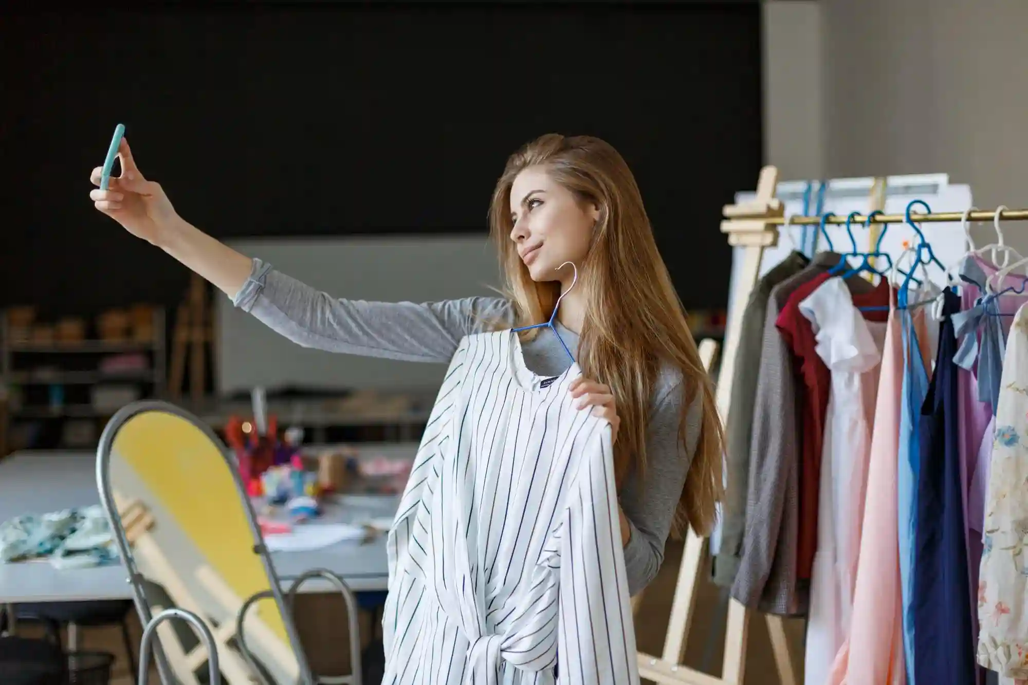 Young blonde woman holding up a striped garment and taking a selfie video in a fashion studio.