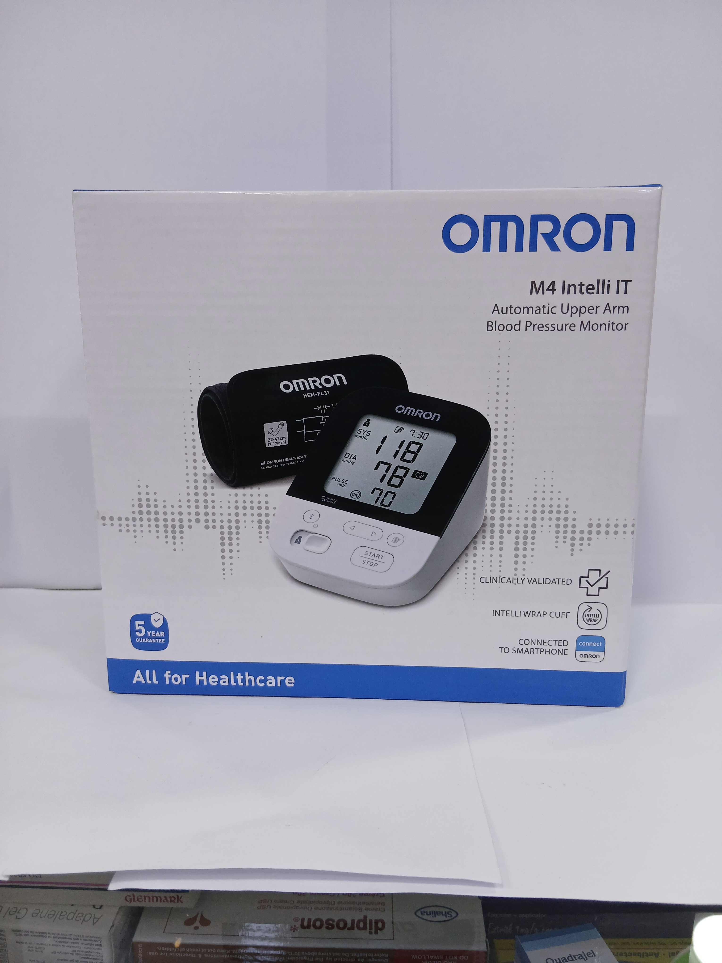 White cardboard packaging displays a blood pressure monitor with an attached black upper arm cuff, featuring a digital screen showing readings, placed against a plain background.