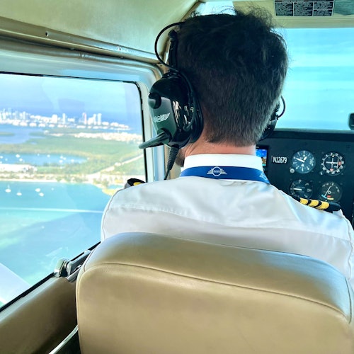 飞行员戴着耳机驾驶飞机，通过驾驶舱窗户可以看到海岸线和城市景观。