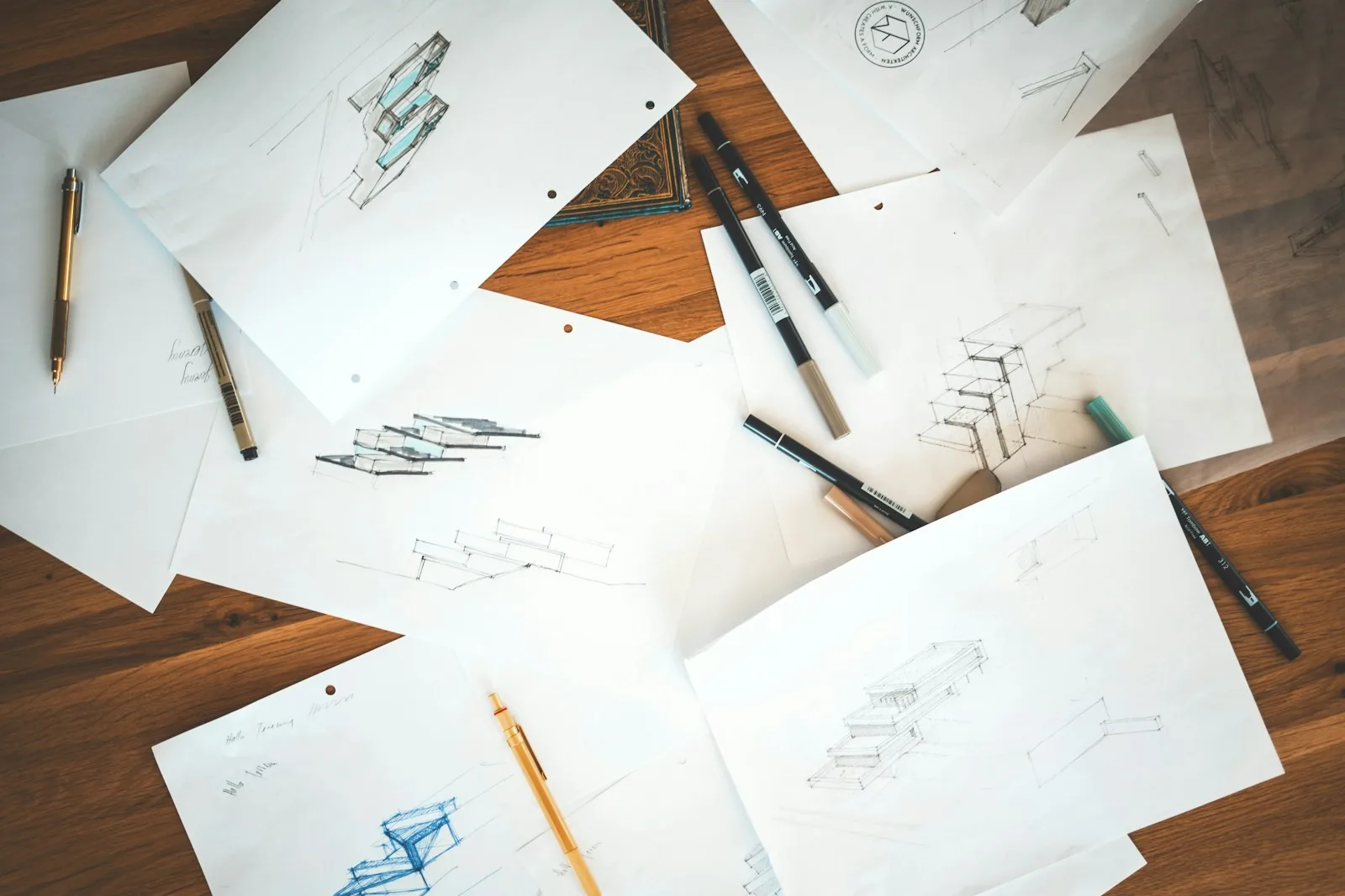 Pencil sketches of stacked architectural forms on loose sheets of paper scattered across a wooden desk with pens and markers