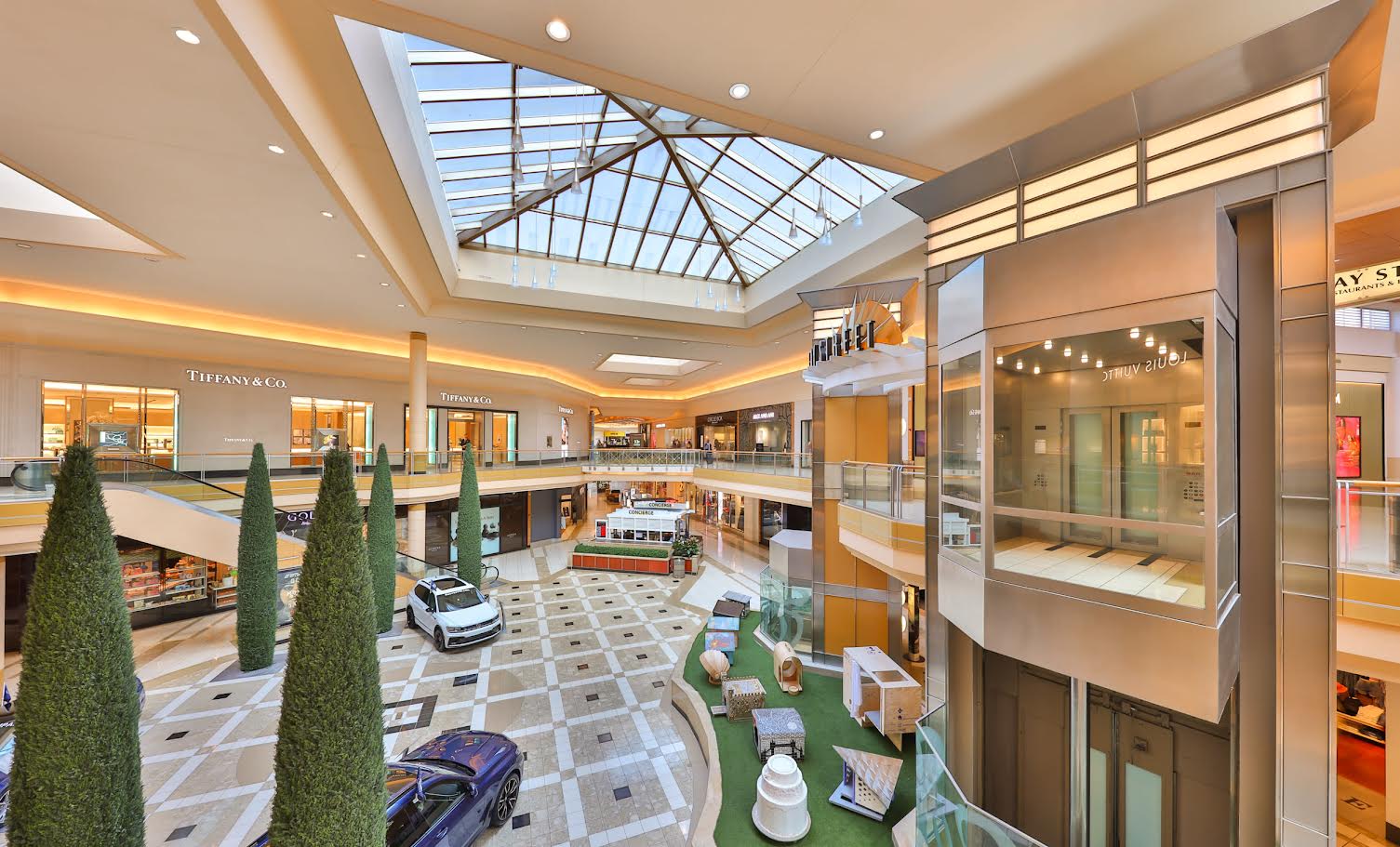 Rendering of International Plaza's renovated interior showing a spacious shopping corridor with large skylights, warm lighting, retail storefronts, decorative topiaries, and polished flooring