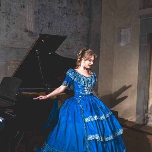 一位身穿蓝色连衣裙的女士站在一架三角钢琴旁，置身于一间灯光昏暗、墙面富有纹理的房间中。