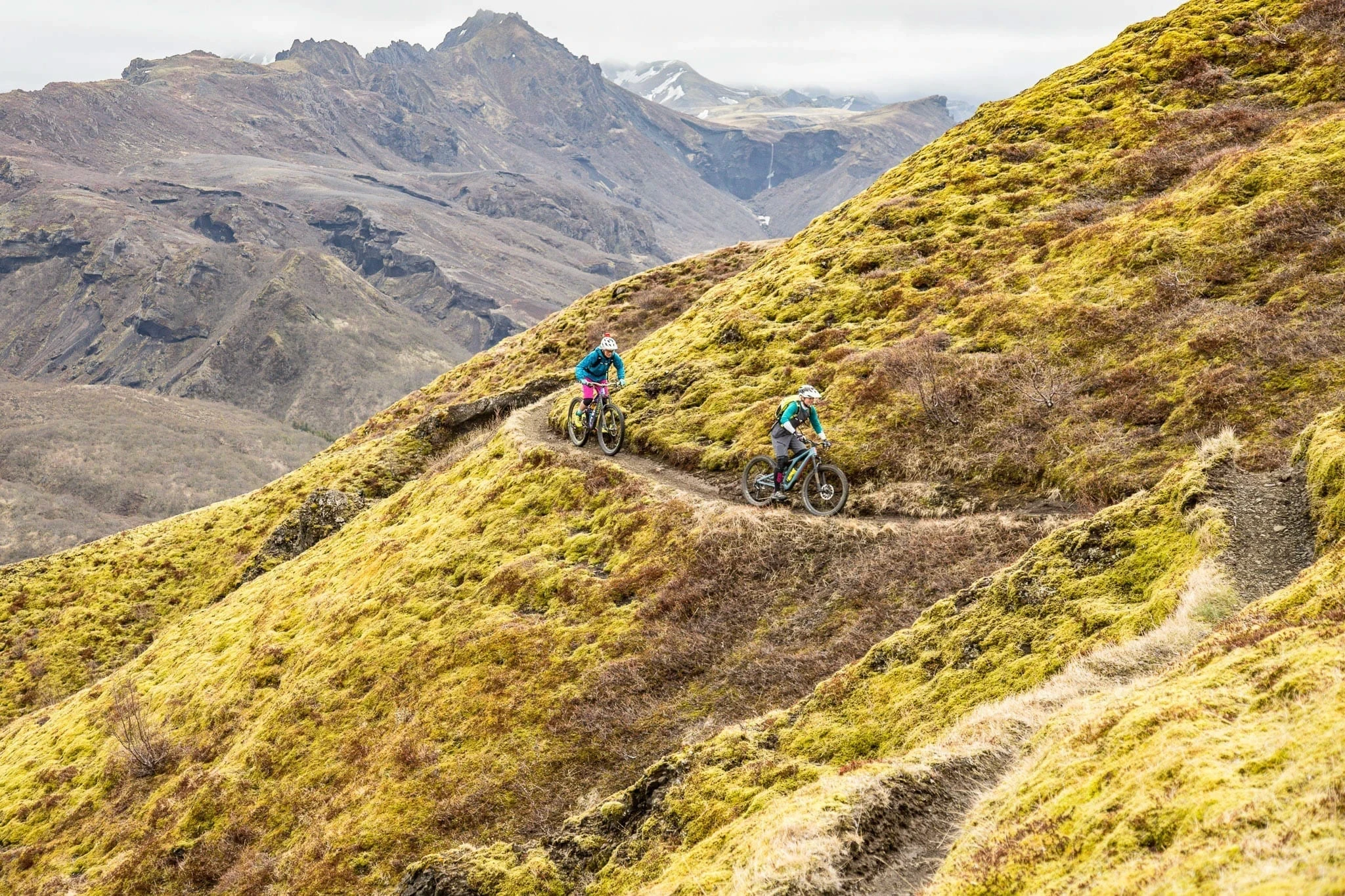 Two mountain bikers riding singletrack across a mossy slope with rocky mountains.