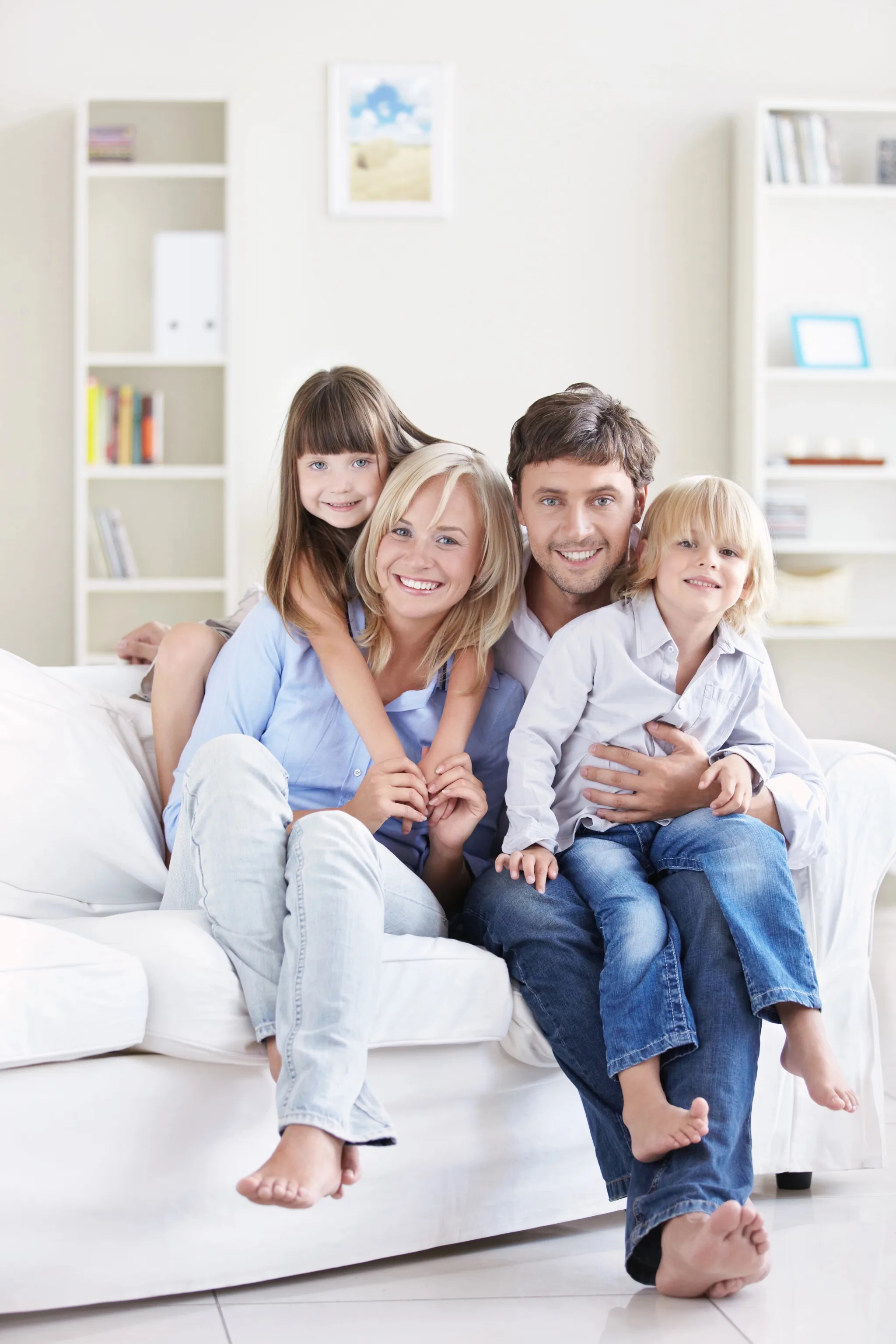 A happy family of four—two parents and two children—sitting together on a white couch in a bright, minimalist living room with white shelving units in the background.