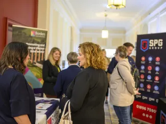 People are engaging at a business networking event in a hallway. Stands display promotional banners and materials. The atmosphere is professional and lively.