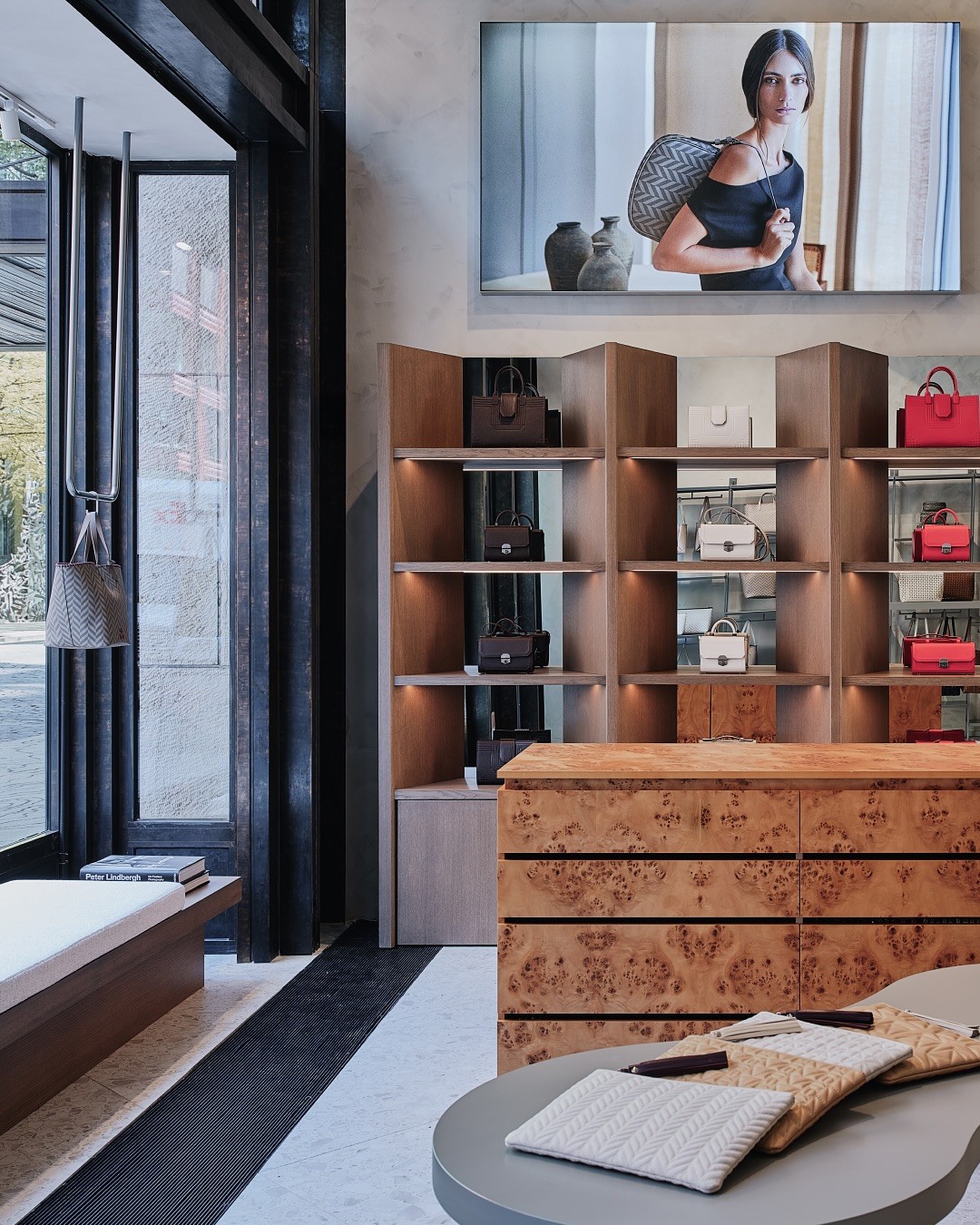 Luxury handbag boutique interior by Misela featuring sculptural display tables, burl wood cabinetry, mirrored walls, and shelves of red and neutral leather handbags in a warm, minimalist retail space.