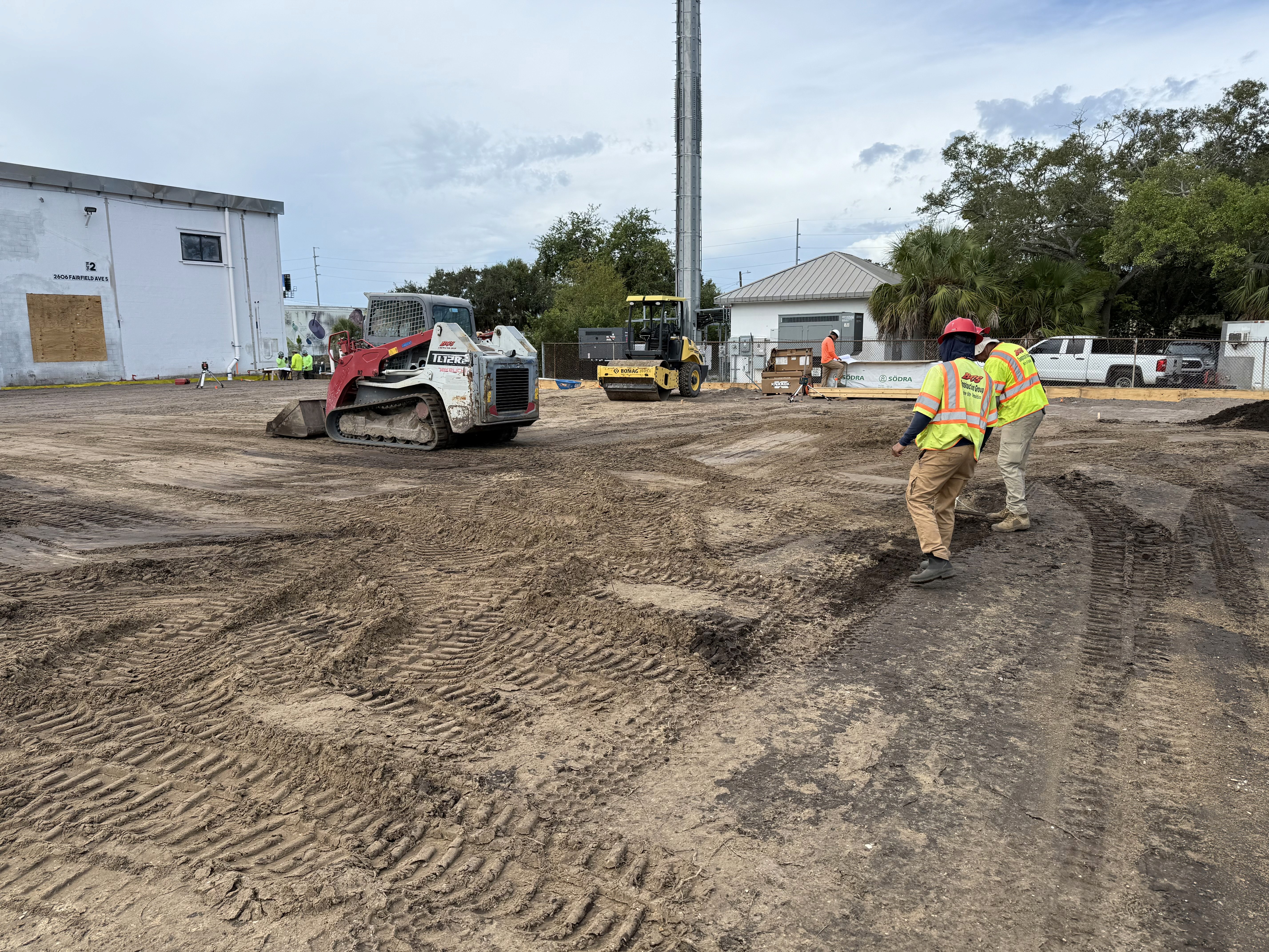 Construction workers in safety vests and helmets preparing a dirt site with heavy equipment, including a skid steer loader and roller compactor