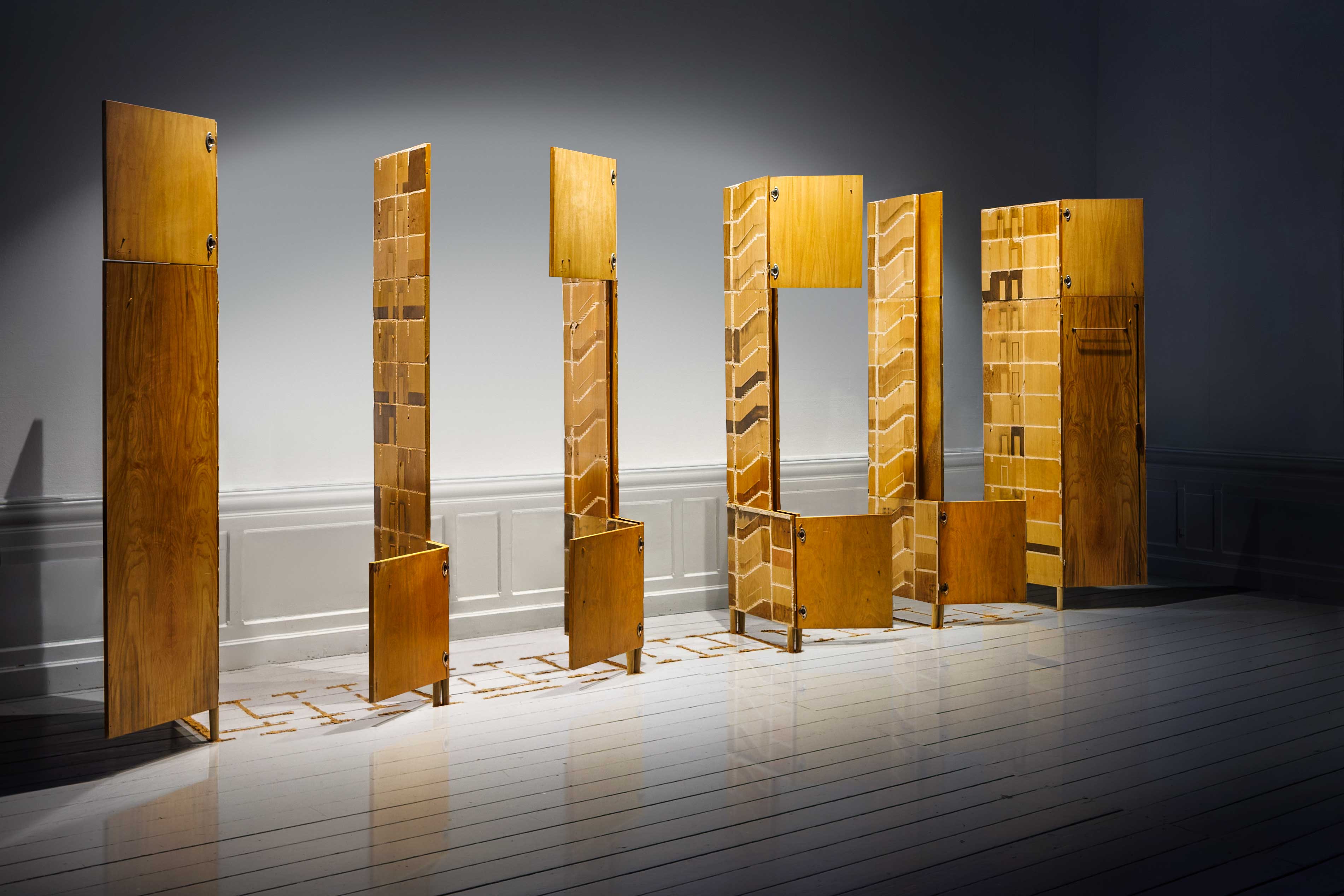 Angled view of multiple vertical wooden cabinet structures with open compartments, displayed on a white wooden gallery floor.