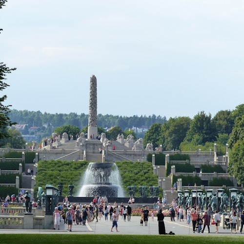 Большой открытый парк с центральным монолитом, фонтаном на переднем плане и множеством гуляющих людей. Территория окружена деревьями и зеленью.