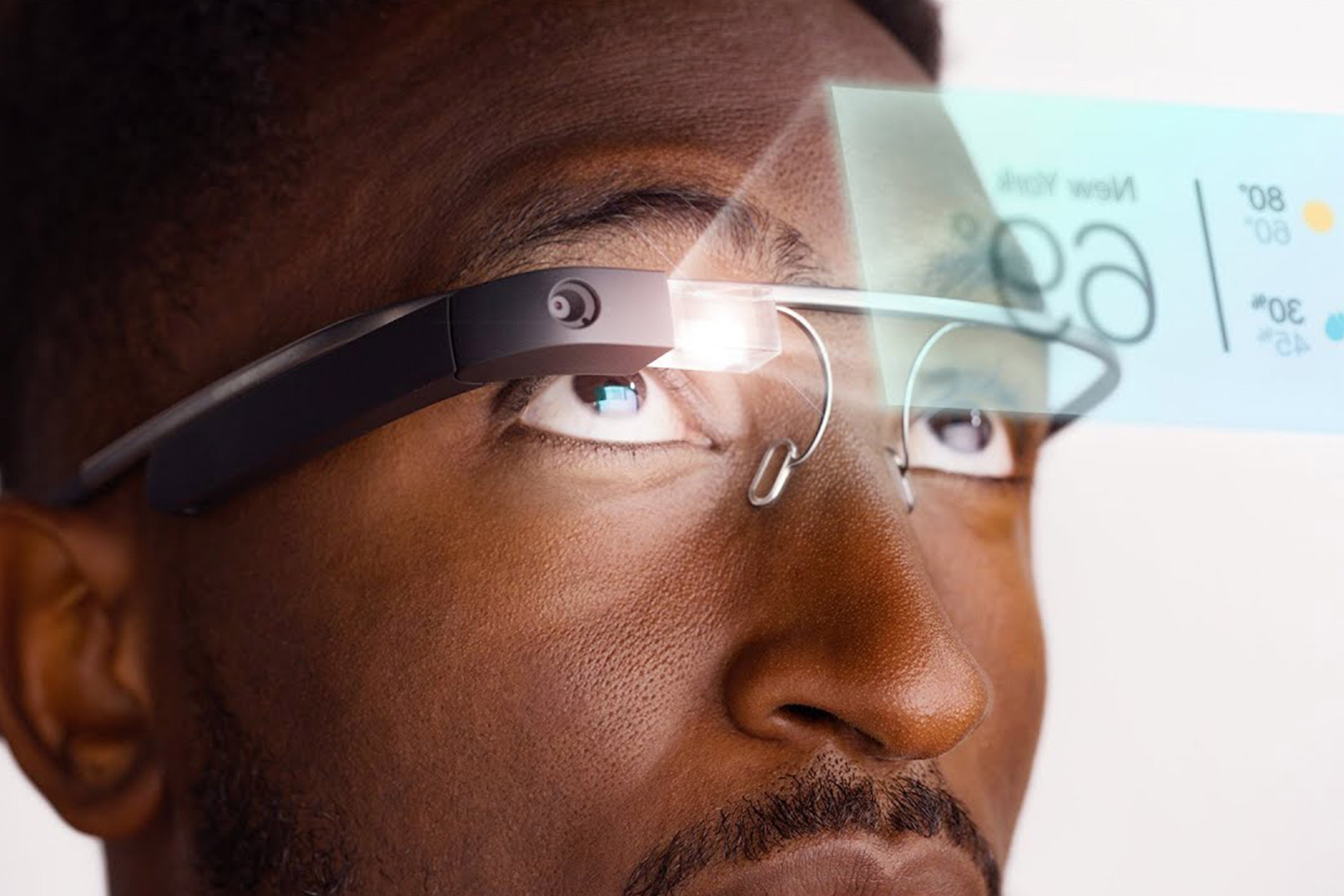 A close-up of man wearing Google Glass and looking up while an user interface screen is superimposed mid-air.