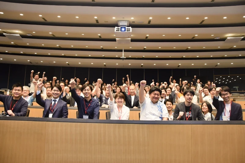Group photo of smiling attendees standing and raising their fists in celebration inside a large auditorium.
