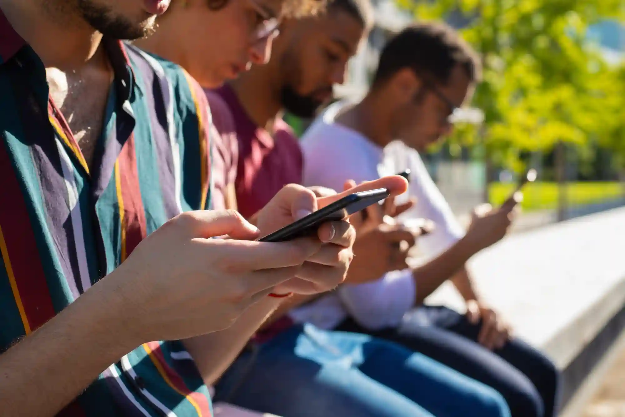 Close-up of diverse young people sitting outdoors, all engrossed in looking at their smartphones.