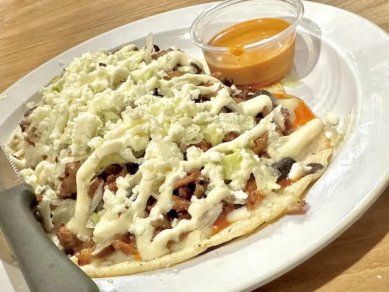 A delicious open-faced taco, topped with melted cheese, fresh onions, and savory meat, sits on a white plate with a small cup of spicy orange dipping sauce, showcasing the vibrant flavors found at Oneco Farmers Market, Bradenton, FL.