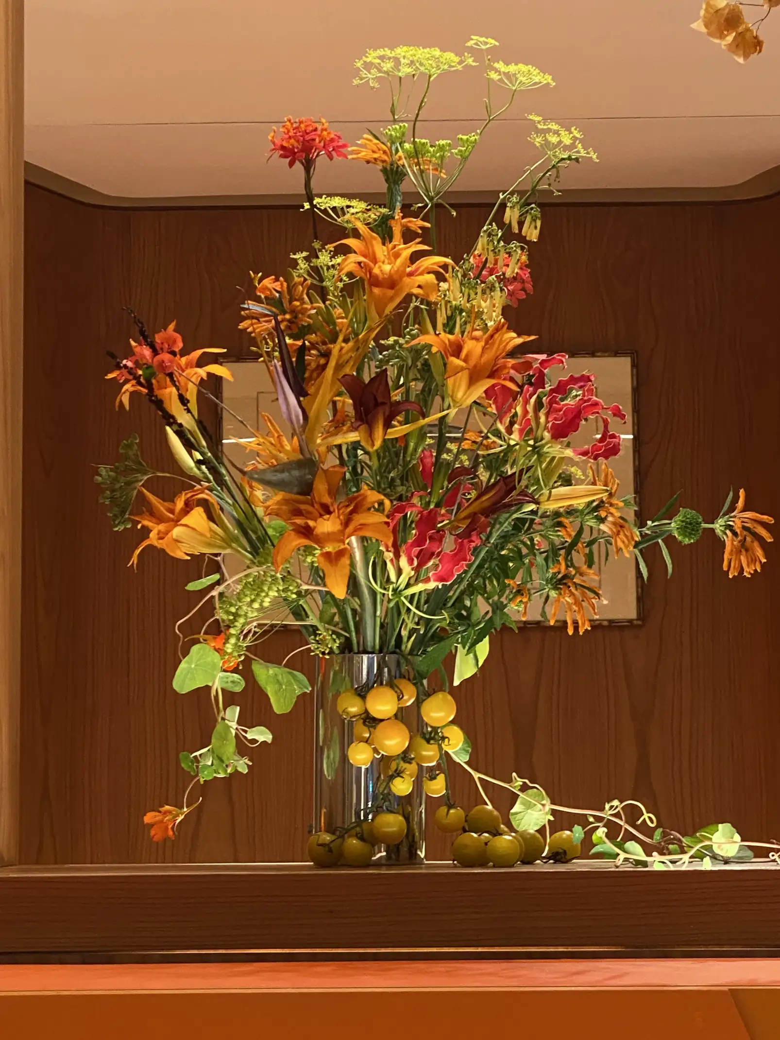 Arreglo floral exuberante en jarrón metálico con base de pequeños tomates amarillos. Las flores, en tonos cálidos de naranja, rojo y amarillo, se combinan con follaje verde y ramas delicadas, destacando sobre un fondo de madera cálida que realza la composición luminosa y orgánica.