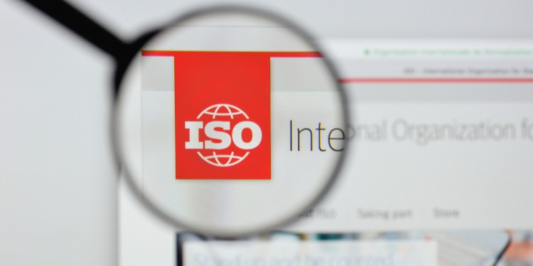 The image shows a magnifying glass focused on the red and white logo of ISO (International Organization for Standardization), emphasizing its presence on a computer screen, suggesting a focus on standards and quality control.