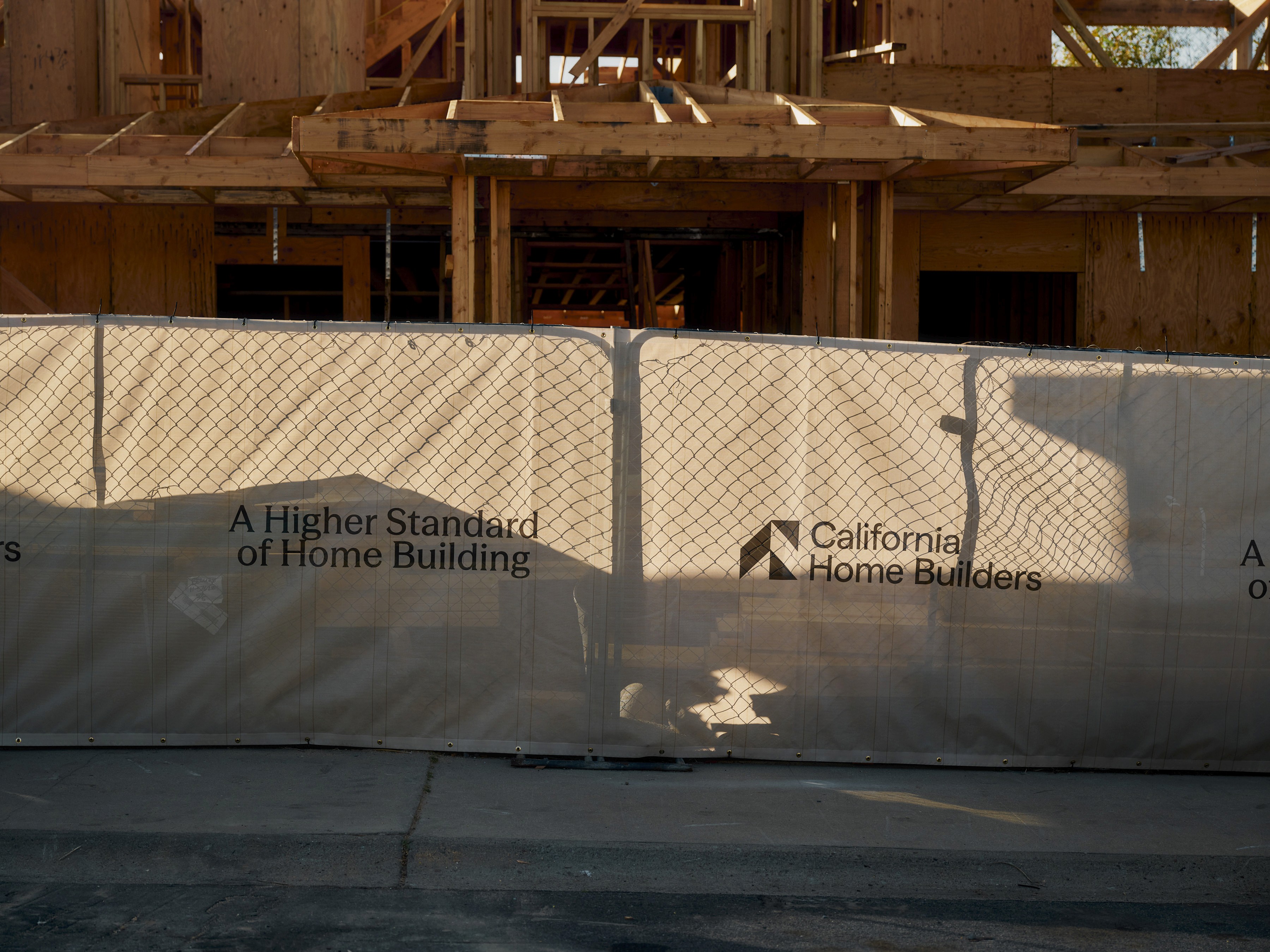 Close-up of a construction fence screen displaying the text: 'A Higher Standard of Home Building' next to the 'California Home Builders' logo. A two-story wooden house frame is visible in the background