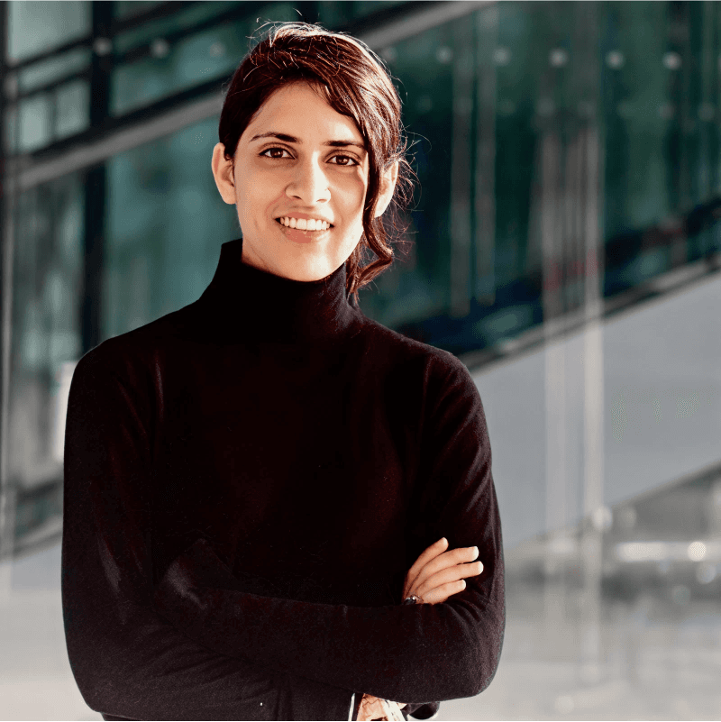 A confident woman in a black turtleneck stands with arms crossed, smiling in a modern indoor setting.