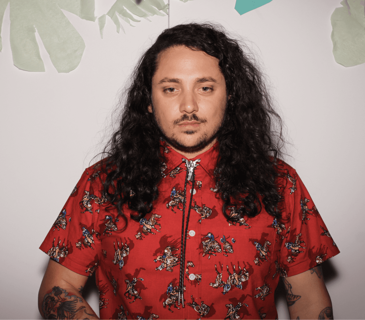 Director Anthony Falvo, who specializes in comedy, stands against a white background decorated with paper cutouts. He wears a bold red shirt patterned with cowboy illustrations and a bolo tie, his long curly hair and tattoos adding to his distinctive, playful presence.