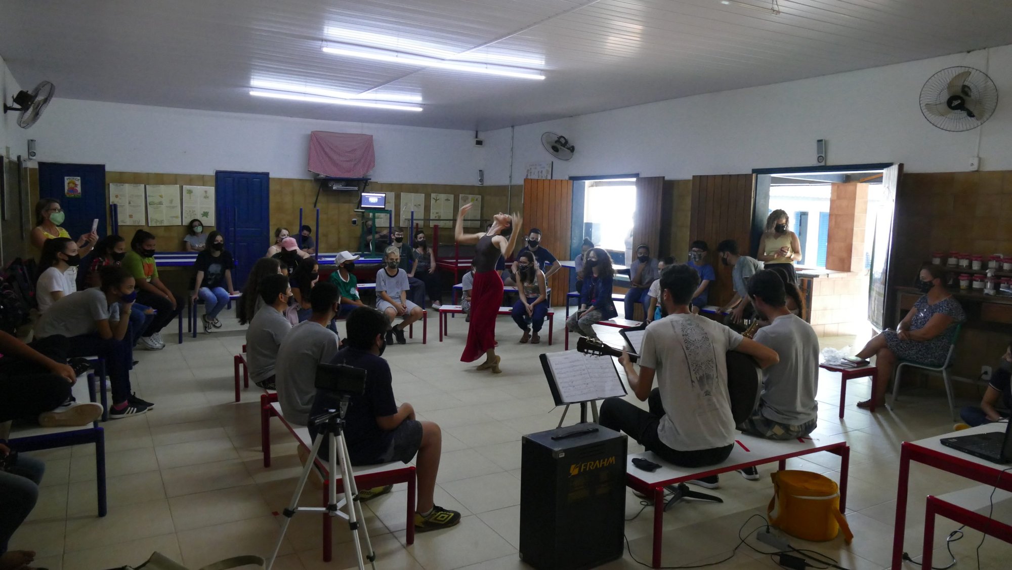 Bailarina dança entre alunos em salão de escola, acompanhada por músicos