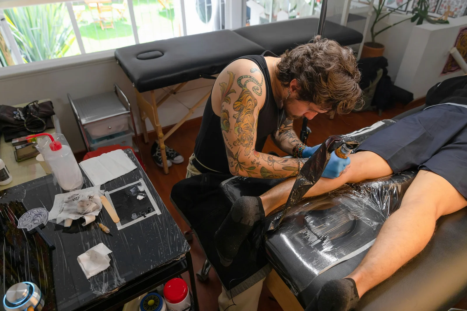 Tattoo artist with visible sleeve tattoos working on client's arm in studio setting with equipment and supplies.