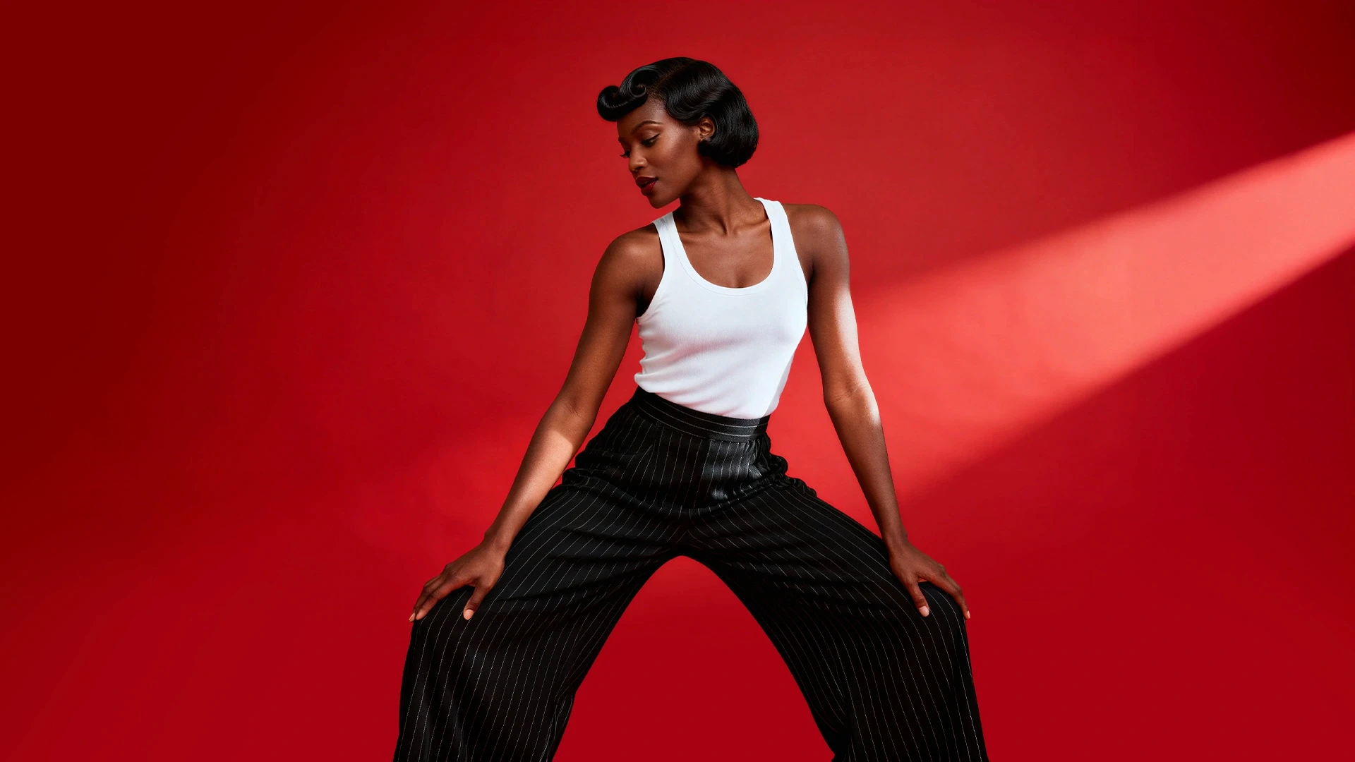 A stylish woman poses dynamically in a white tank top and black pinstriped pants against a bold red background.