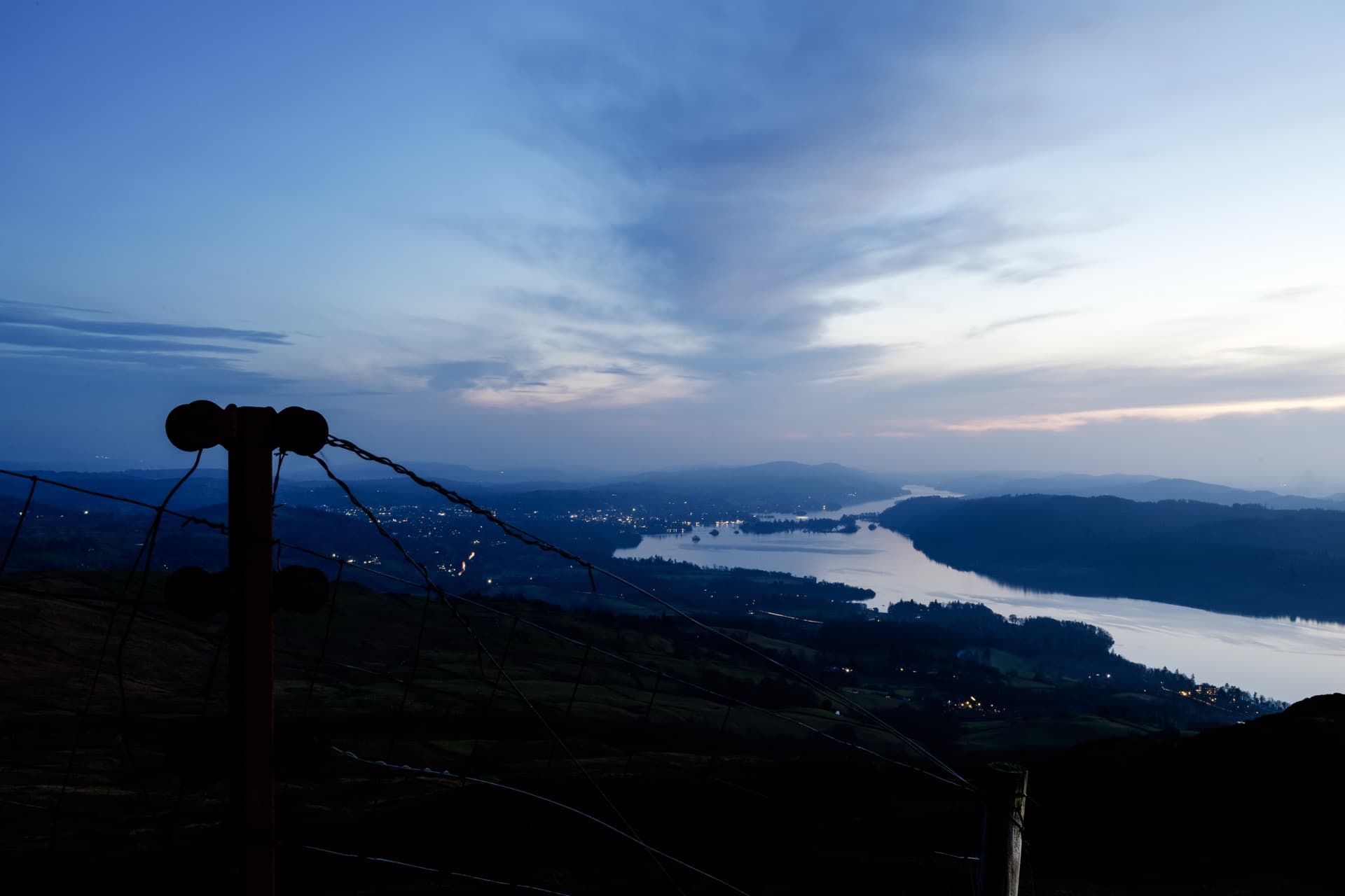 Wire fence post silhouetted at left against blue hour sky, Windermere stretching through valley below with scattered settlement lights emerging