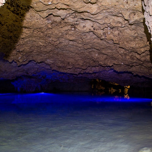 Caverna subterrânea com teto rochoso, iluminada por uma luz azul refletindo na água cristalina abaixo.