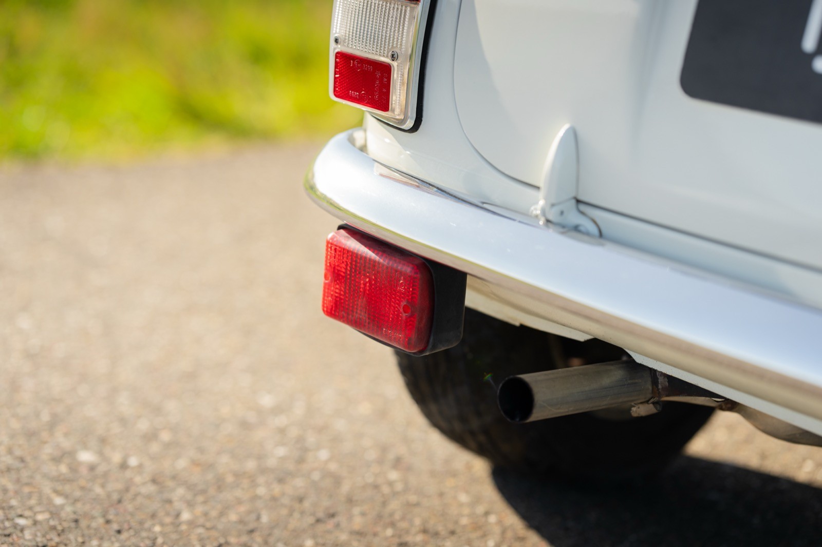 Close-up of the rear of a white vehicle, featuring a red taillight on a paved road with grass in the background.