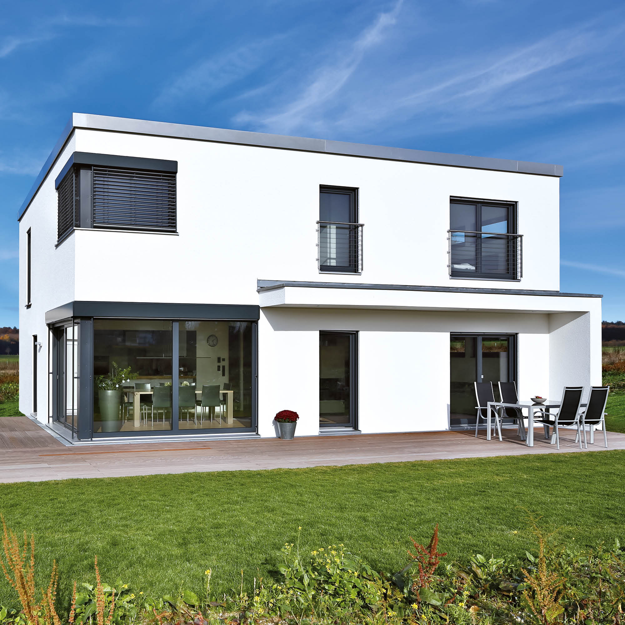 Modernes weißes zweistöckiges Haus mit großen Fenstern, Balkonen und einem Sitzbereich auf der Terrasse.