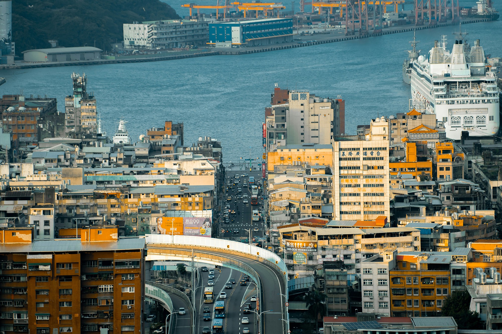 這張圖像展示了基隆市區的景色，前景是密集的城市建築和一條高架道路，背景是基隆港口和停泊的郵輪。圖像呈現了基隆作為港口城市的繁忙和現代化特點。攝影者為 YEH CHE WEI。