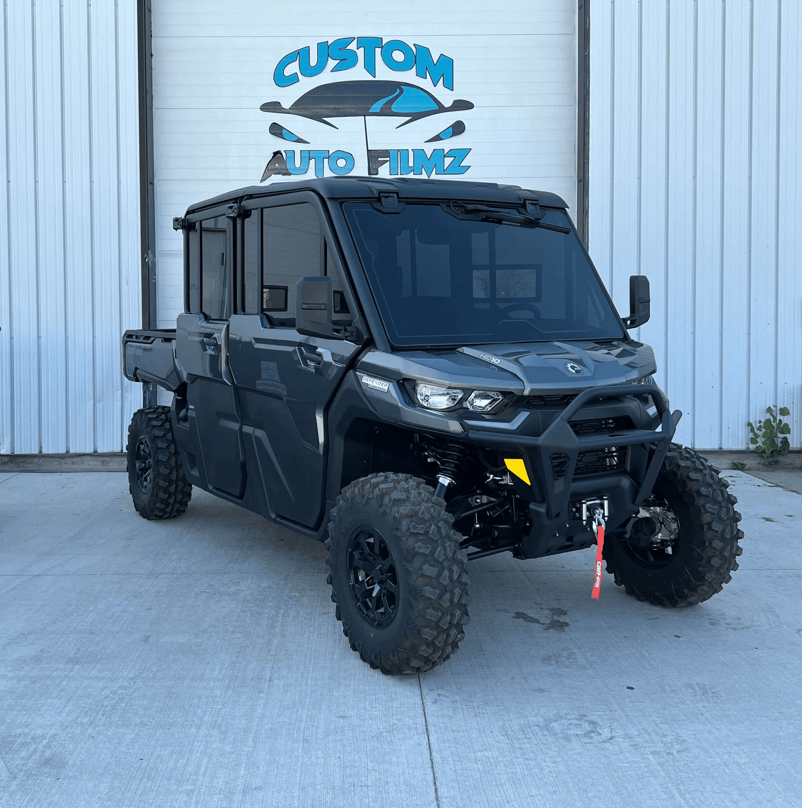A black Can-Am Defender side-by-side utility vehicle is parked on a concrete driveway in front of a white industrial building.