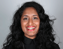 A woman with long, dark, curly hair is captured in a close-up portrait against a solid light gray background. She is wearing a black top with a collar, and her face is lit up with a broad smile, revealing her teeth. Her eyes are dark, and her eyebrows are dark and well-defined. The image focuses solely on her face and upper body, highlighting her expression and attire. No other objects or text are visible in the frame.