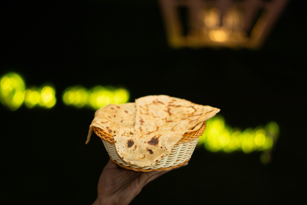 A close-up of a large, hearty chicken wrap or shawarma, filled with meat and fries, generously drizzled with white and orange sauces and sprinkled with parsley, served on a wooden cutting board with a side of French fries.