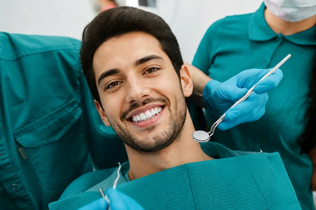 Dentist examining a smiling male patient's teeth using professional dental tools in a clinic.